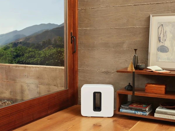 Interior shot of a white speaker on a wooden floor, with a window showing a mountain view and a wooden shelf with objects.