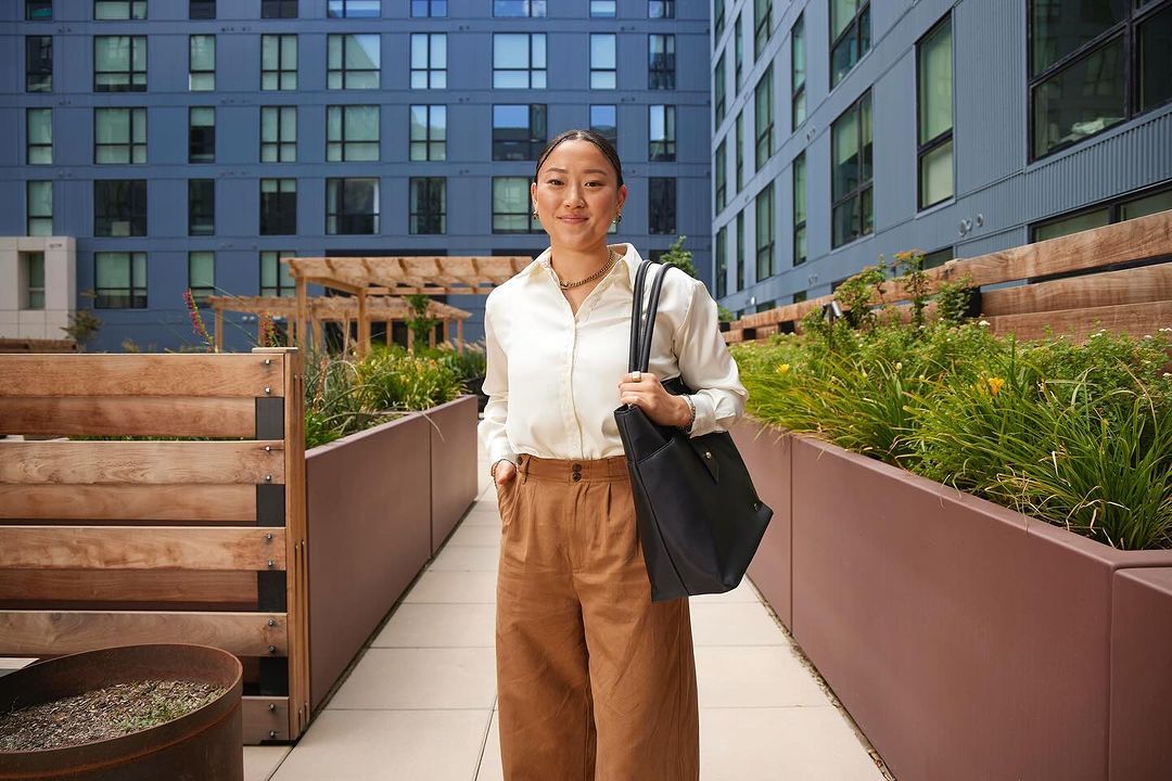 A fashionable Asian woman stands outside a modern apartment building. She's wearing a white shirt, brown pants, and carrying a black bag.