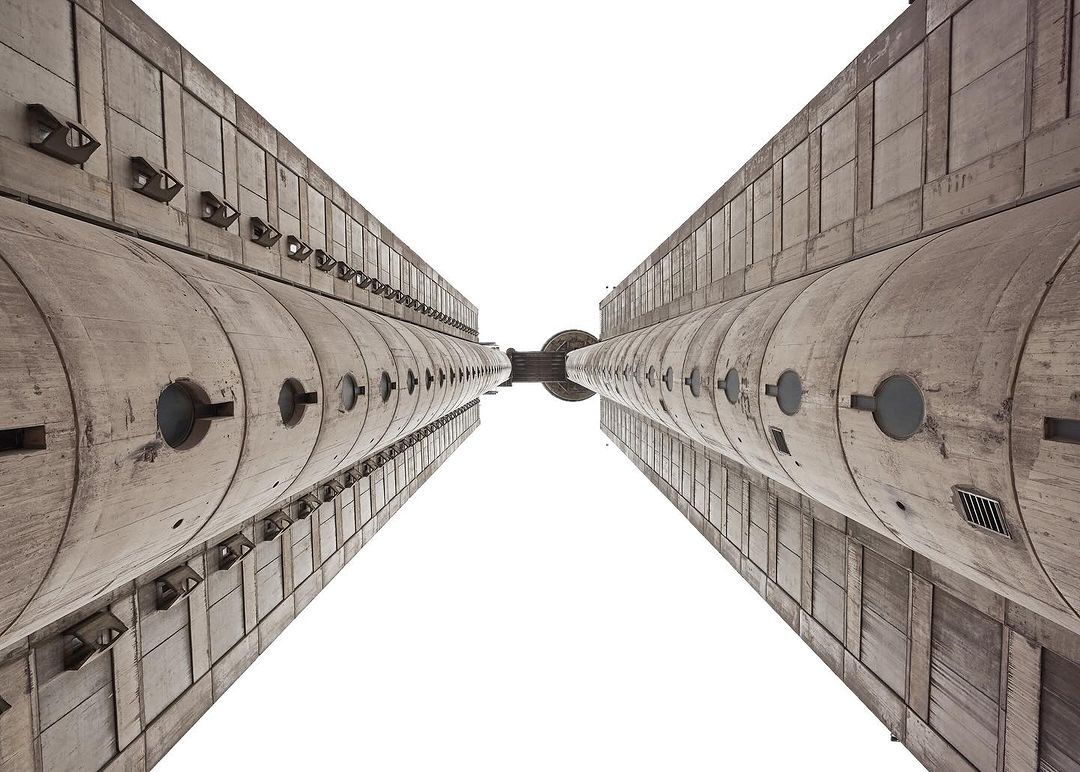 Worm's eye view of two massive Brutalist concrete towers converging against a bright white sky, showcasing raw architectural grandeur.