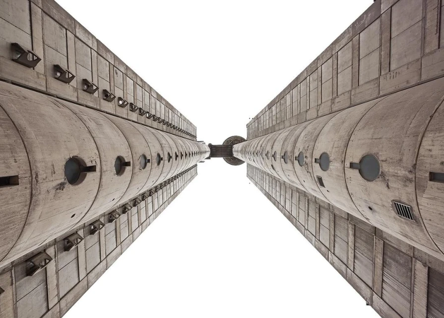 Worm's eye view of two massive Brutalist concrete towers converging against a bright white sky, showcasing raw architectural grandeur.