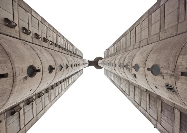 Worm's eye view of two massive Brutalist concrete towers converging against a bright white sky, showcasing raw architectural grandeur.