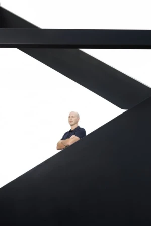 Bald adult man in a polo shirt standing with arms crossed in an indoor setting framed by a black architectural structure.