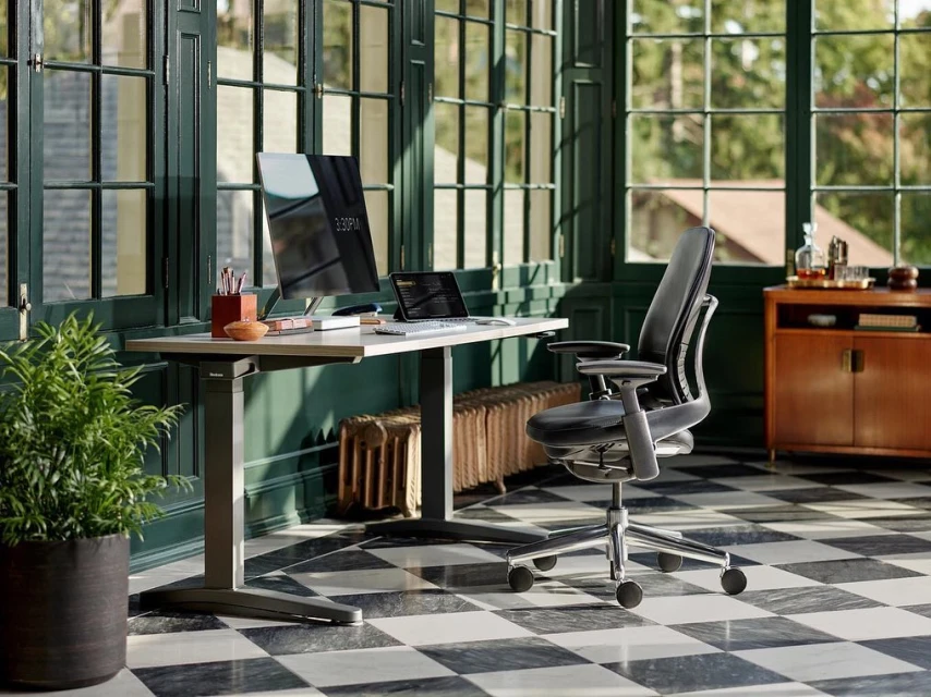 Modern office setup features a sleek desk with a computer and laptop, a stylish black office chair, and natural light from large windows.