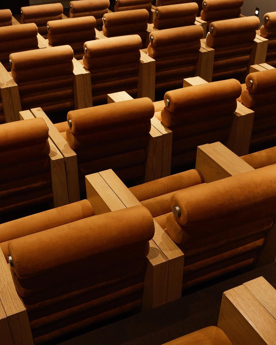 Rows of tan-colored, upholstered seating with wooden frames in a spacious auditorium create a modern and comfortable viewing experience.