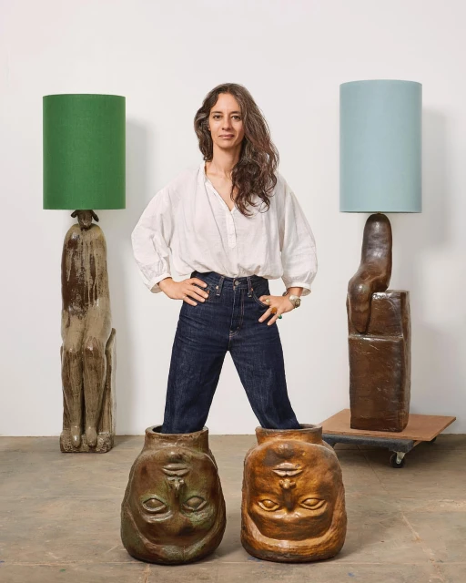 Adult woman stands between lamps with shades and ceramic bases, and ceramic face vases in a bright, indoor photo studio setting.