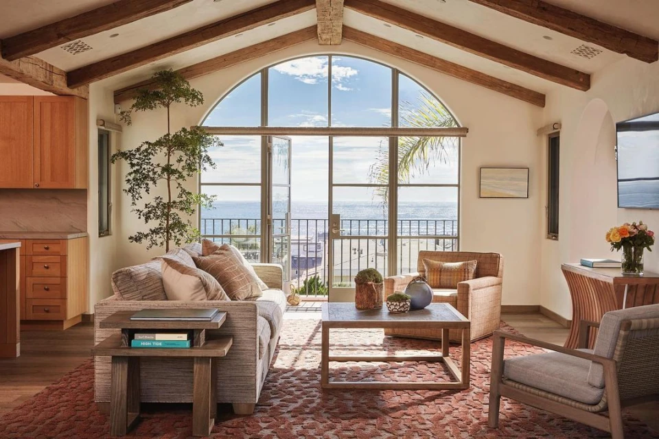 Cozy living room with natural light, featuring a grey couch, wooden coffee table, and a view of the ocean through large windows.