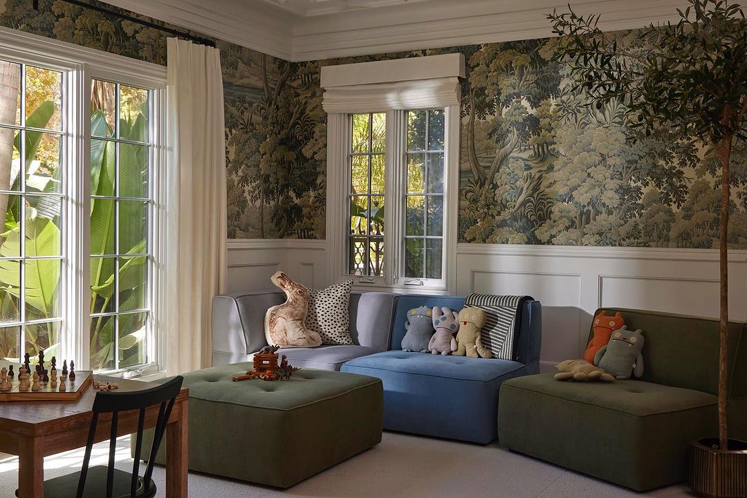Cozy living room interior with neutral-toned sofas, plush pillows, and decorative wallpaper featuring lush trees and calming natural light.