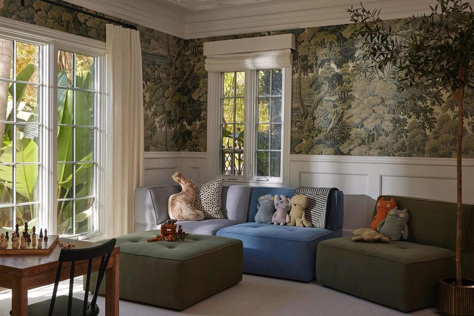 Cozy living room interior with neutral-toned sofas, plush pillows, and decorative wallpaper featuring lush trees and calming natural light.