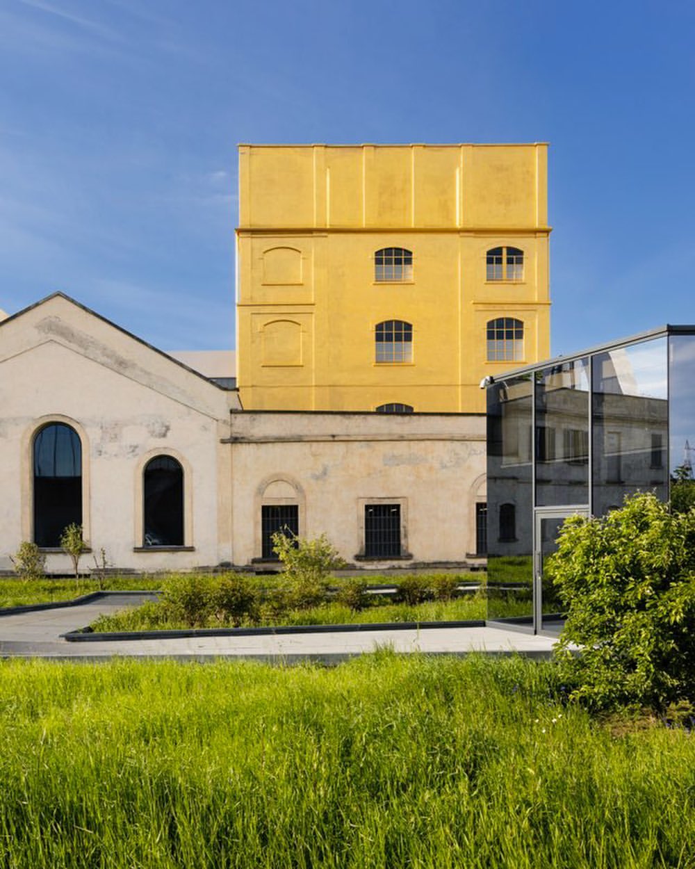 Architectural shot of a modern building complex featuring a golden tower, green space, and a glass structure under a clear blue sky.