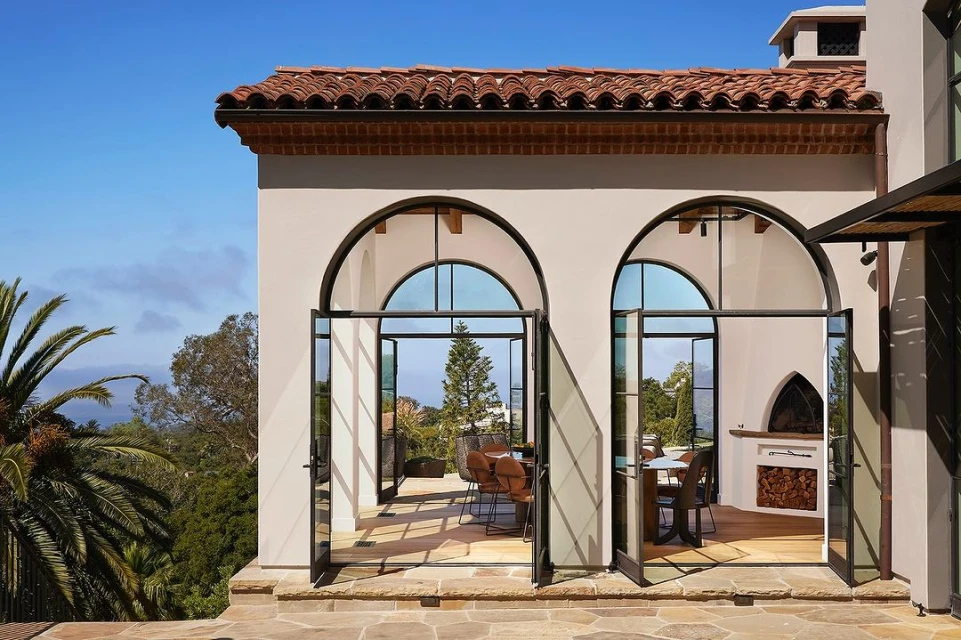 Exterior view of a luxury house with an open terrace, featuring stone floors, arched windows, and an ocean view with lush trees.