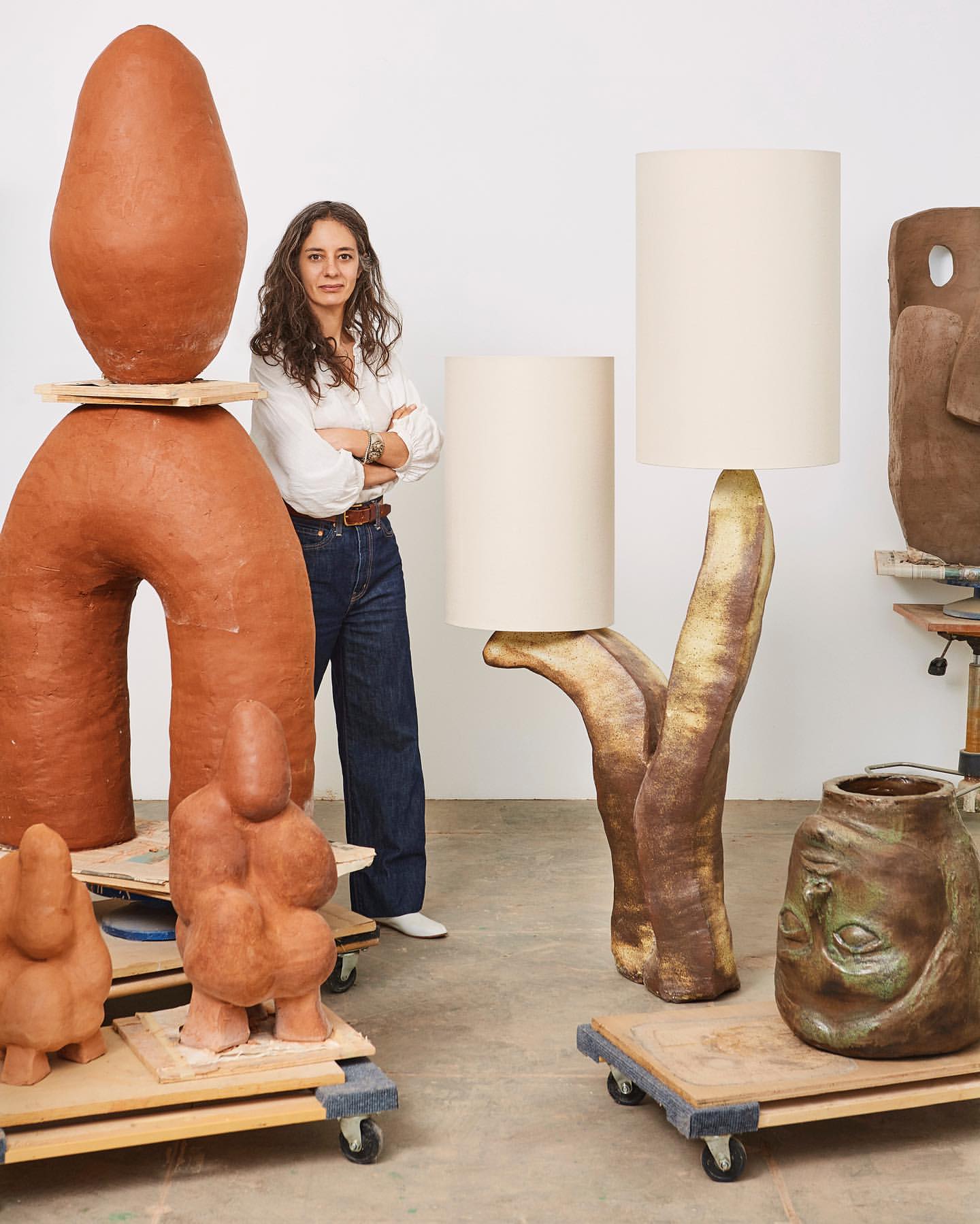 Female artist poses in studio with handcrafted clay lamps and sculptures in neutral colors, set against a white backdrop.