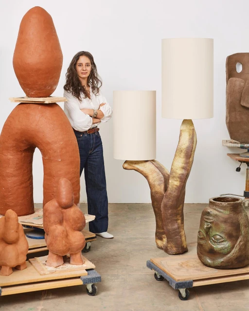 Female artist poses in studio with handcrafted clay lamps and sculptures in neutral colors, set against a white backdrop.