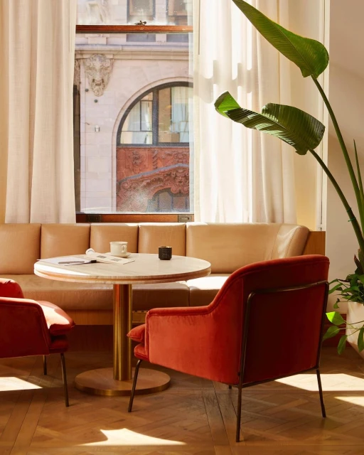 Indoor cafe seating area with a round table, two red velvet chairs, and a green plant near a window showcasing an urban building.