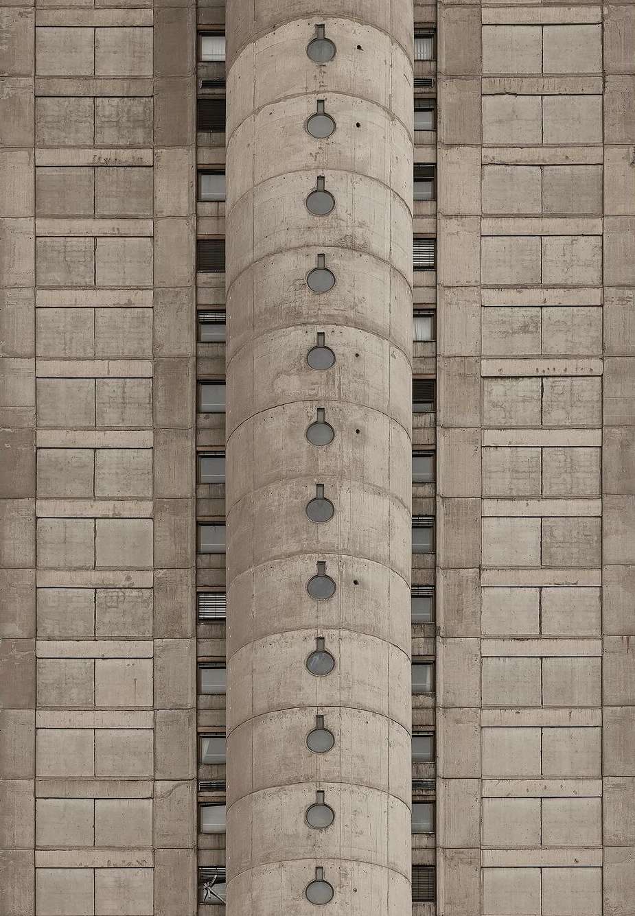 Concrete building facade with repeating geometric pattern and cylindrical tower featuring small, round windows running vertically.