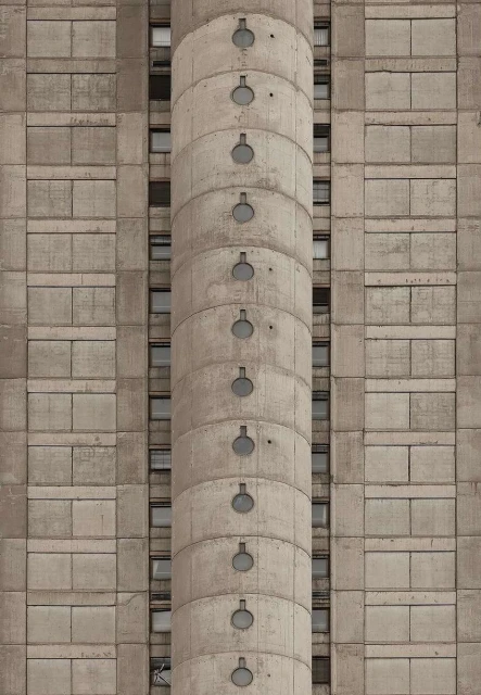 Concrete building facade with repeating geometric pattern and cylindrical tower featuring small, round windows running vertically.