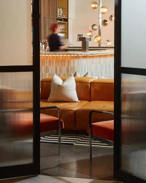 Interior view of a hotel lobby lounge with tan leather sofa, red chairs, globe lighting, and a bar area with a blurred figure.