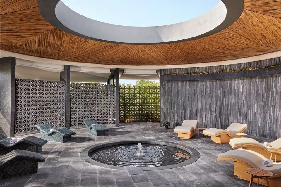 Open-air spa area with tiled floor, textured walls, circular water feature, and cushioned lounge chairs under a circular bamboo ceiling.