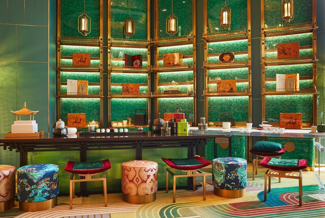 Interior of a tea room featuring a tea counter with stools, decorative objects, and a green display wall with shelves and lighting.