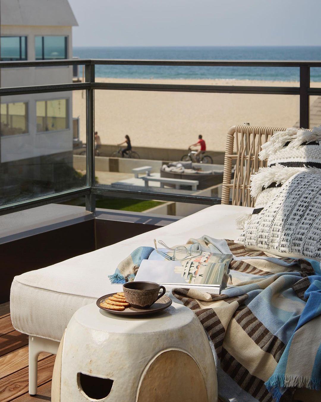 Outdoor lounge area on a sunny balcony with a daybed, pillows, blankets, a side table with tea and cookies, and an ocean view.