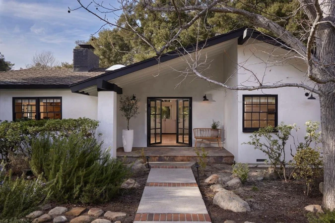 Exterior view of a white one-story house with a black roof, open double doors, and landscaping in the front yard.