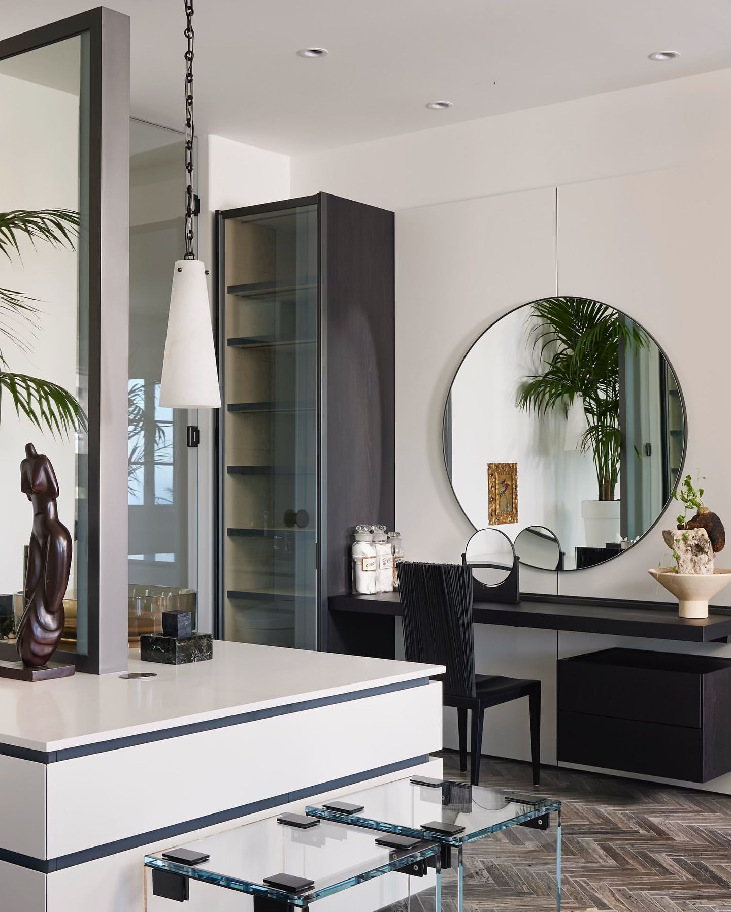 Interior view of a vanity with a round mirror, black chair, and decorative objects, in a room with natural light and neutral tones.