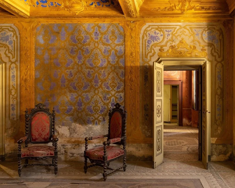 Ornate interior shot of a room with patterned wallpaper, two antique chairs, and a doorway leading to another room in the distance.