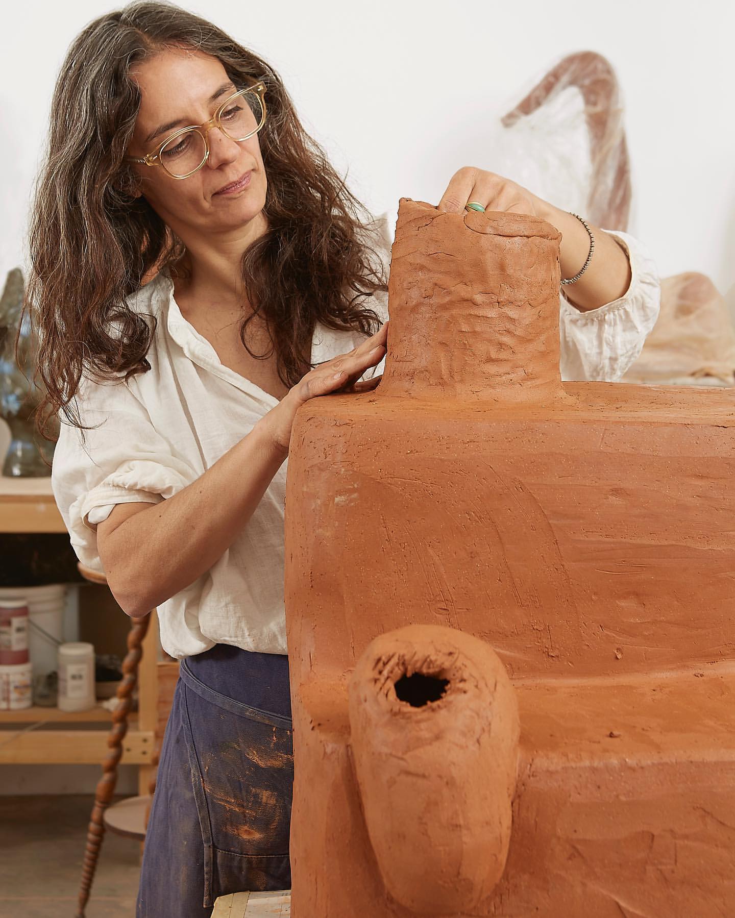 A woman sculpts a large clay form in a studio. The earthy, textured sculpture dominates the frame as the artist shapes its top.