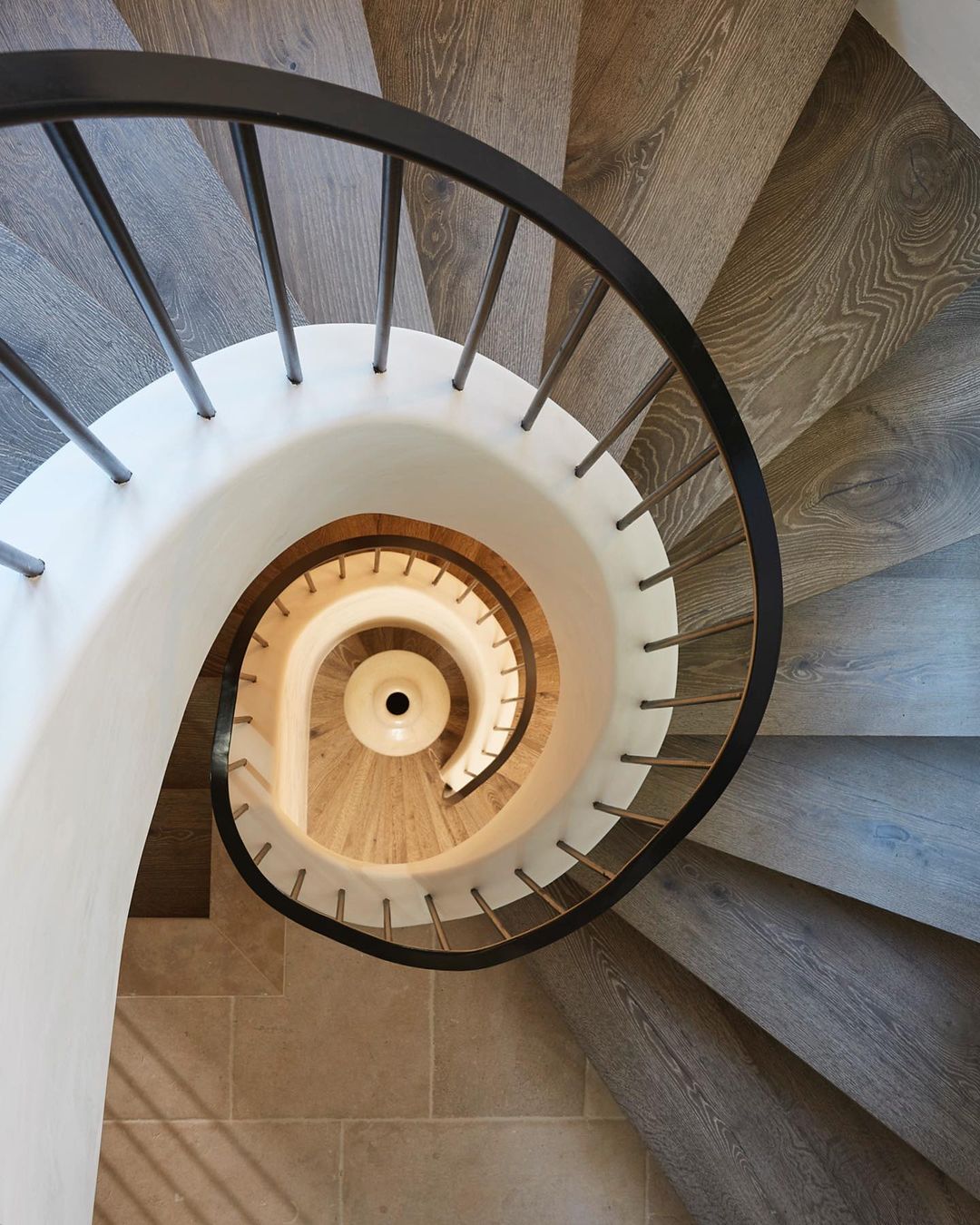 A top-down view of a spiral staircase features light gray wooden steps, a white curved wall, and a black metal railing indoors.