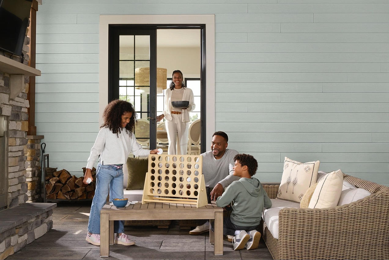 A family of four plays a connect-four game outdoors on a patio. The mother stands in the background by a sliding glass door.