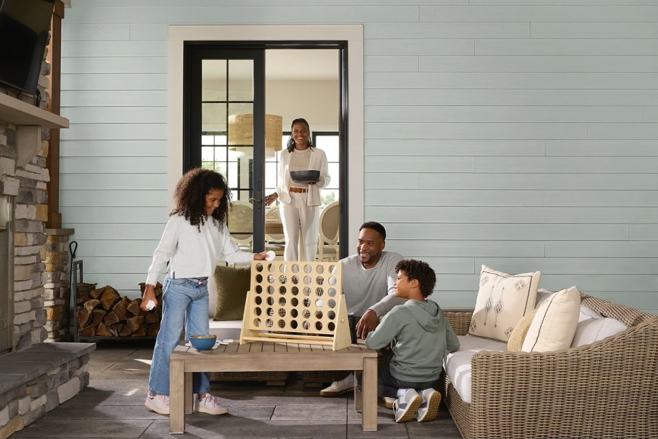 A family of four plays a connect-four game outdoors on a patio. The mother stands in the background by a sliding glass door.