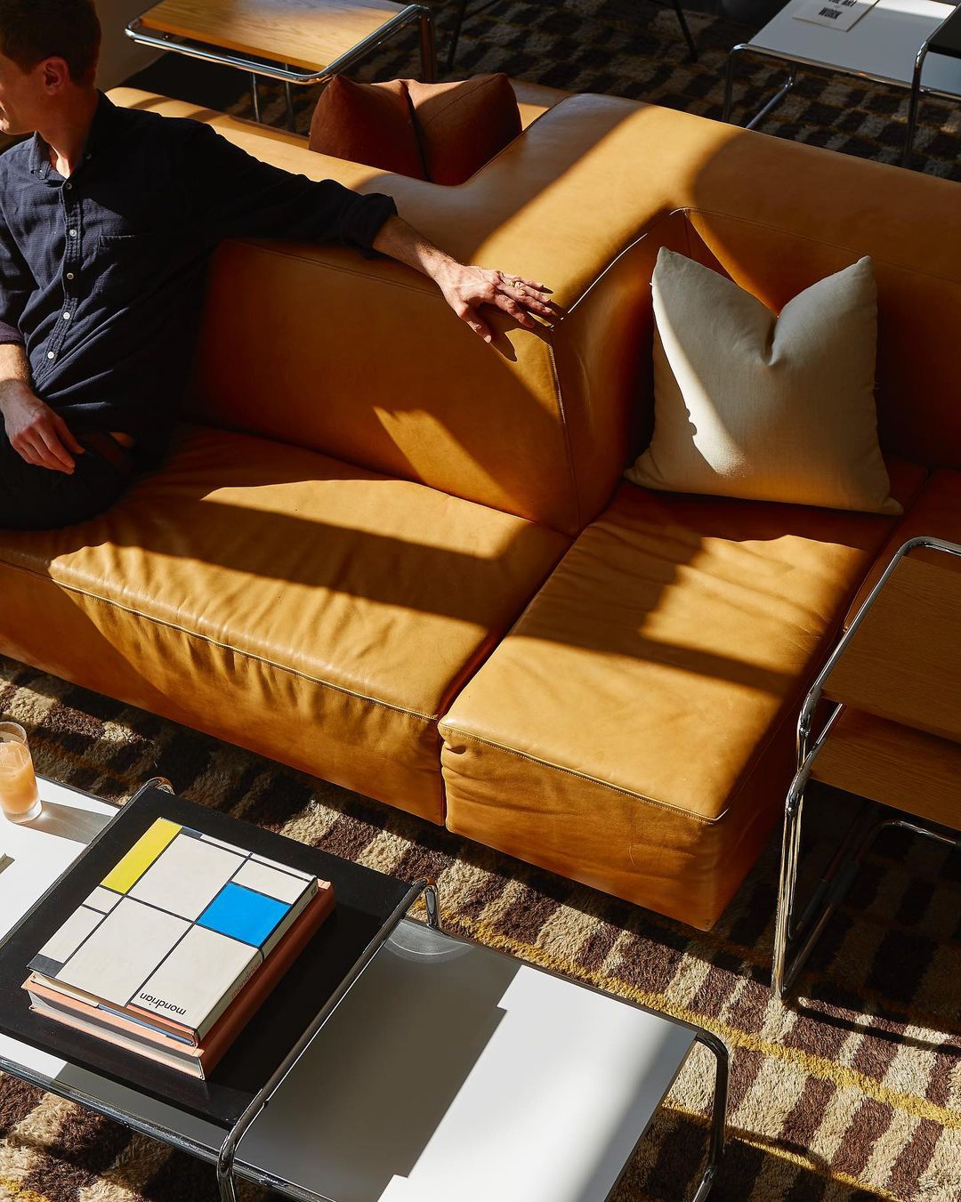 Man sits on a tan leather sectional sofa in a sunlit living room with a modern coffee table and brown patterned rug, interior shot.