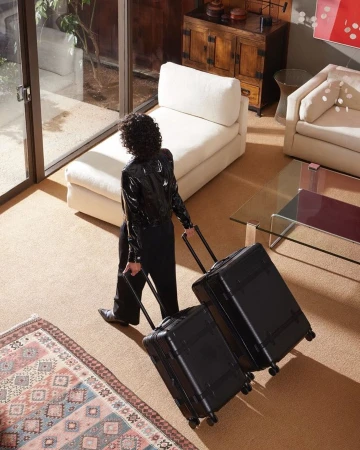 Woman with curly hair, wearing a black jacket, pulls two rolling suitcases through a modern, brightly lit home with neutral decor.