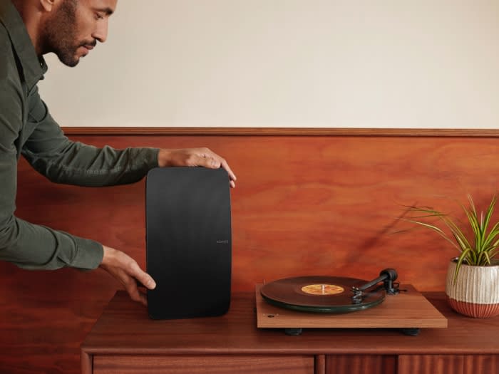 Man holding a black Sonos speaker next to a turntable on a wooden cabinet against a warm-toned wall in an indoor setting.