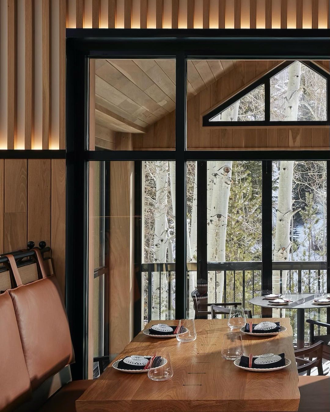 Interior of restaurant dining area with wooden table set for service, bench seating, and a view of aspen trees and snow outside.