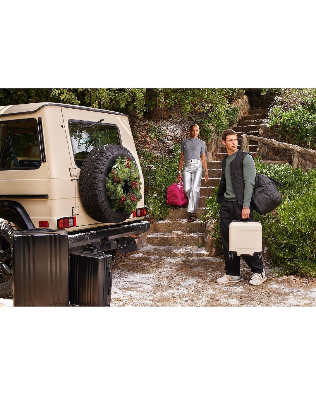 Two models pose with luggage and a luxury SUV in an outdoor setting with greenery and stairs, promoting travel and lifestyle.