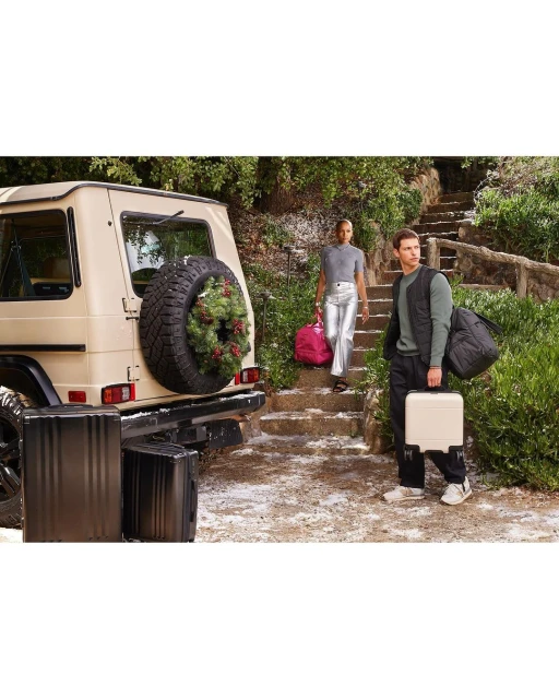 Two models pose with luggage and a luxury SUV in an outdoor setting with greenery and stairs, promoting travel and lifestyle.