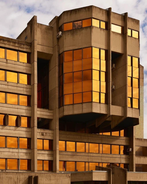 Exterior view of a modern brutalist building featuring a concrete facade and reflective orange tinted windows under a cloudy sky.