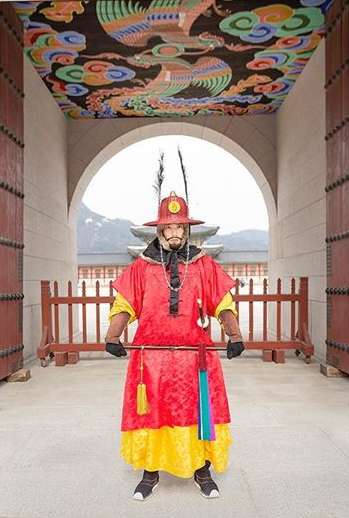 Korean palace guard in traditional red and yellow uniform stands at attention in front of an arched stone gate with painted ceiling.