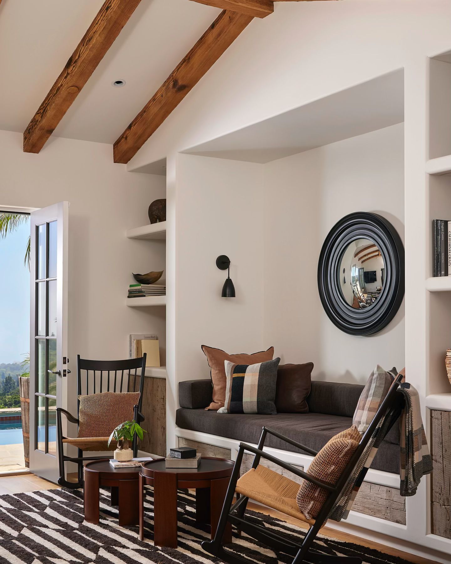 Living room interior with two wooden rocking chairs, a brown sofa, a round mirror, and a black and white striped rug in a bright space.