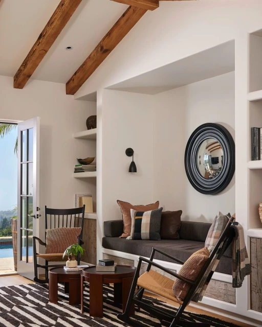 Living room interior with two wooden rocking chairs, a brown sofa, a round mirror, and a black and white striped rug in a bright space.