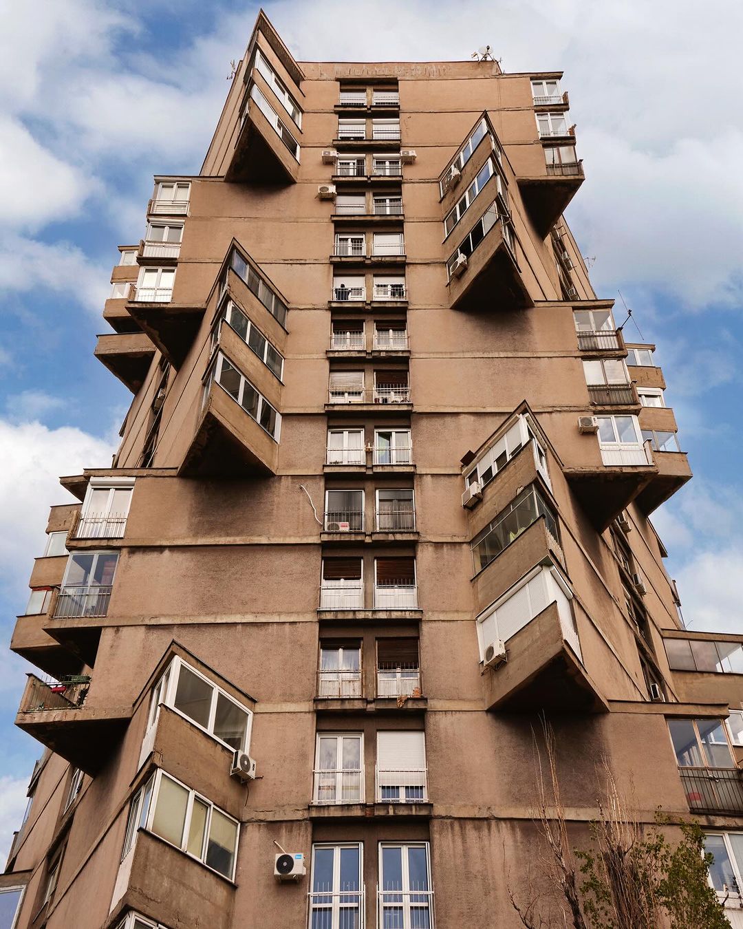 Exterior of a tall, brutalist apartment building with protruding balconies and rows of windows against a blue sky with wispy clouds.
