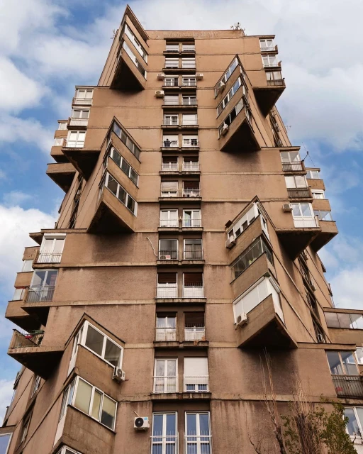 Exterior of a tall, brutalist apartment building with protruding balconies and rows of windows against a blue sky with wispy clouds.