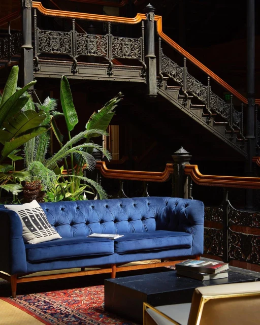Indoor lounge area features a blue velvet couch, coffee table with books, patterned rug, and staircase with ornate iron railing.