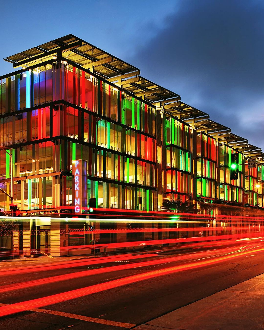 Modern parking garage with colorful facade at night, illuminated by internal lights and light trails from passing traffic.