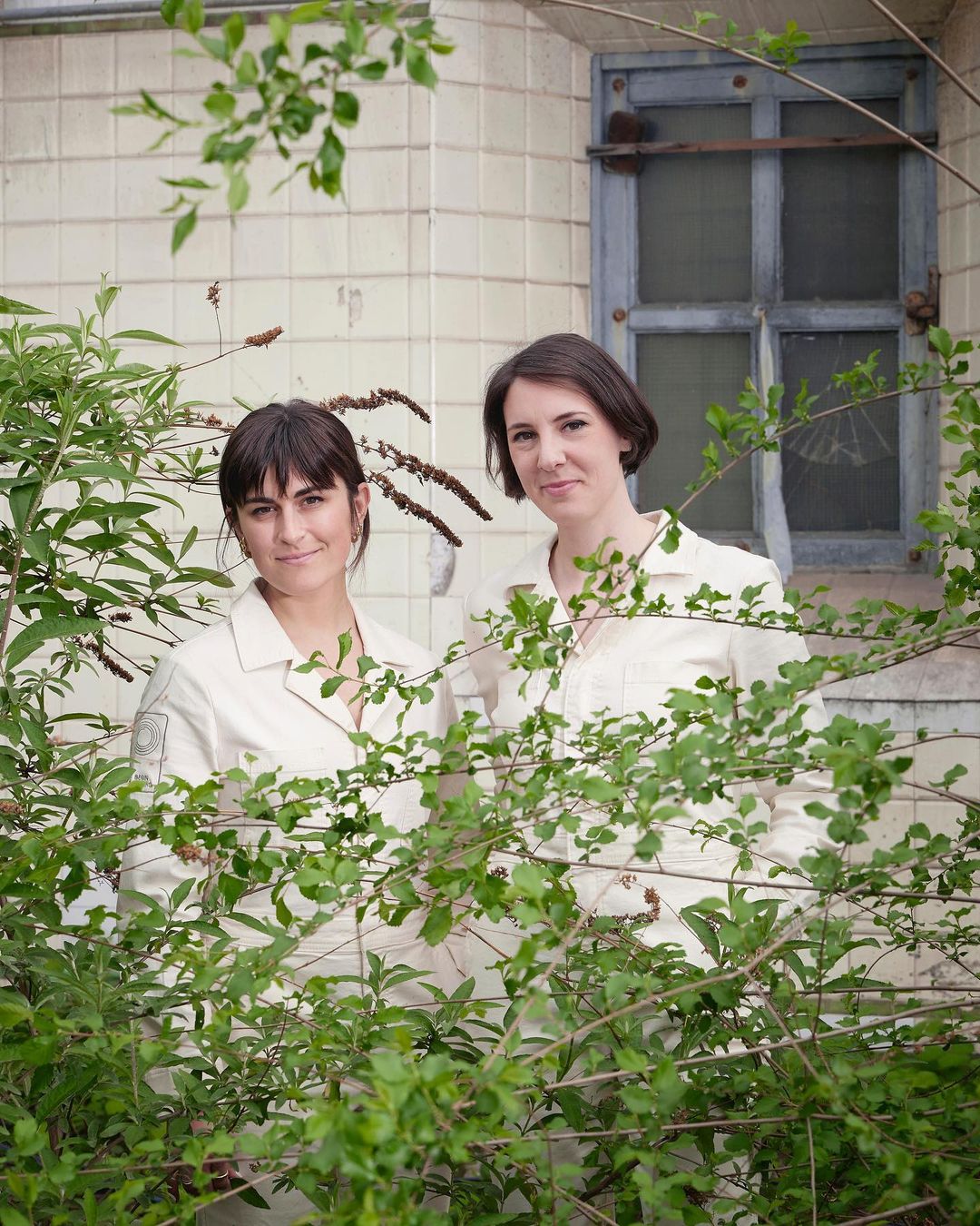 Two women in light-colored outfits stand outside a tile building, framed by green foliage, creating a natural, candid composition.