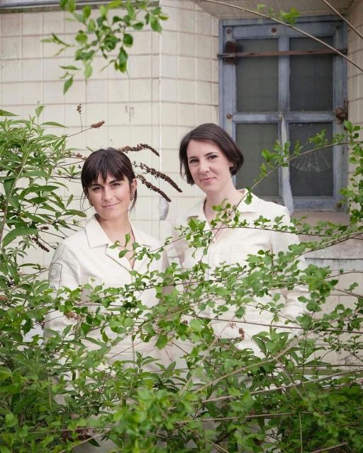 Two women in light-colored outfits stand outside a tile building, framed by green foliage, creating a natural, candid composition.