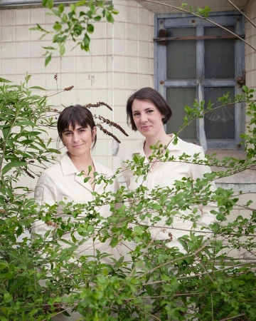 Two women in light-colored outfits stand outside a tile building, framed by green foliage, creating a natural, candid composition.