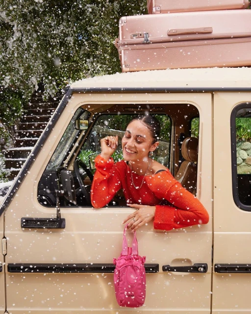 Woman smiling in a car with luggage on top. She's holding a pink bag. Snow is falling around the vehicle, creating a winter travel vibe.