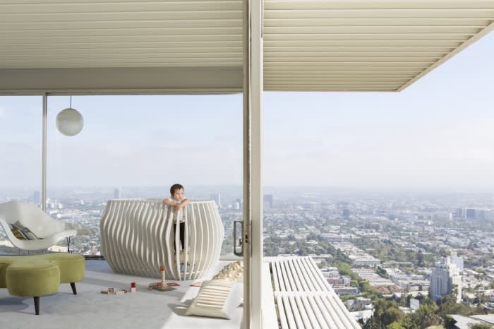 Child in a modern crib on a balcony overlooking a cityscape. The balcony includes modern chairs and stools, and overlooks city views.