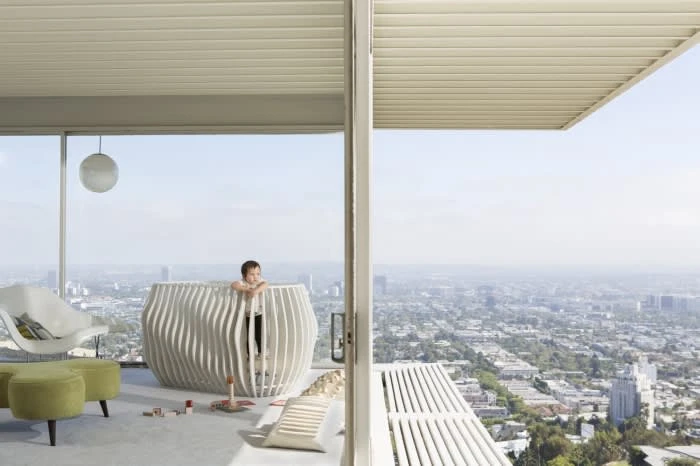 Child in a modern crib on a balcony overlooking a cityscape. The balcony includes modern chairs and stools, and overlooks city views.